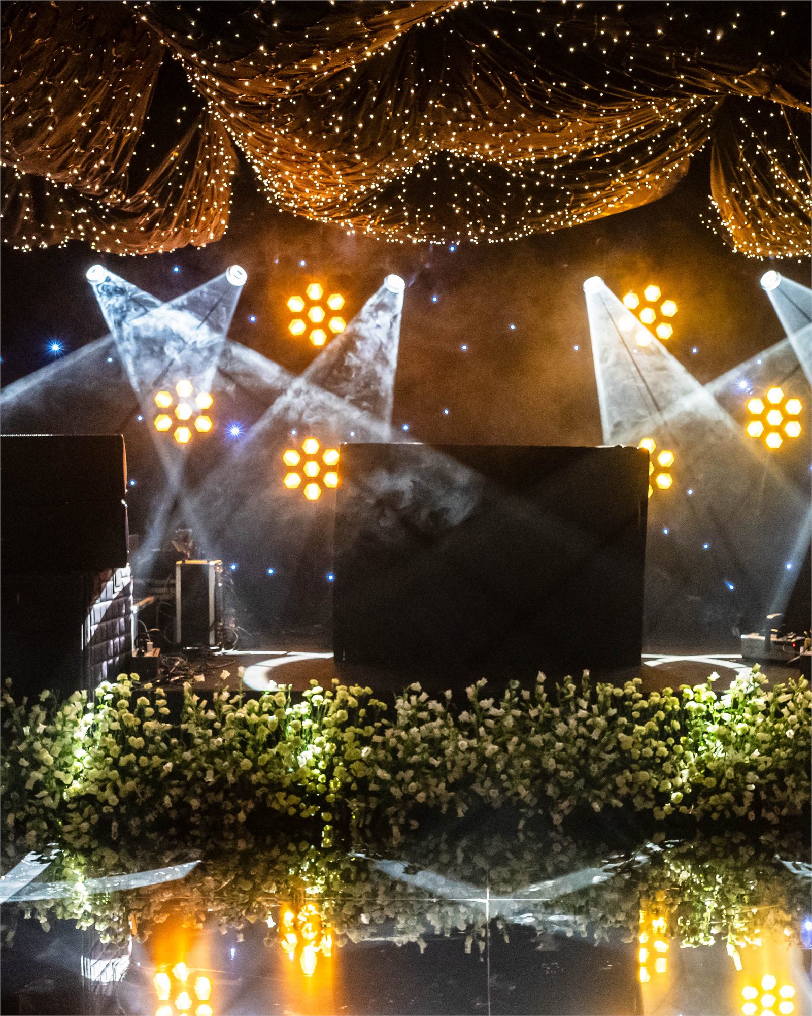A stage under crossed spotlights, banked with white flowers, beneath a canopy of fairy lights
