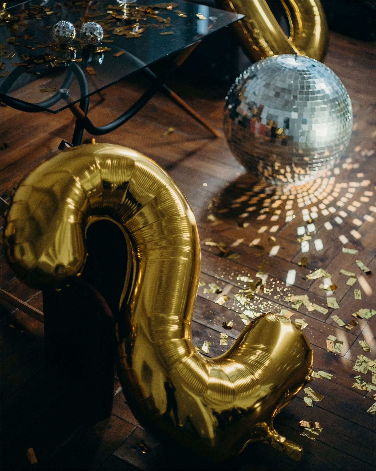 Gold foil balloons and a mirror ball scattering light across a dark wooden floor strewn with confetti