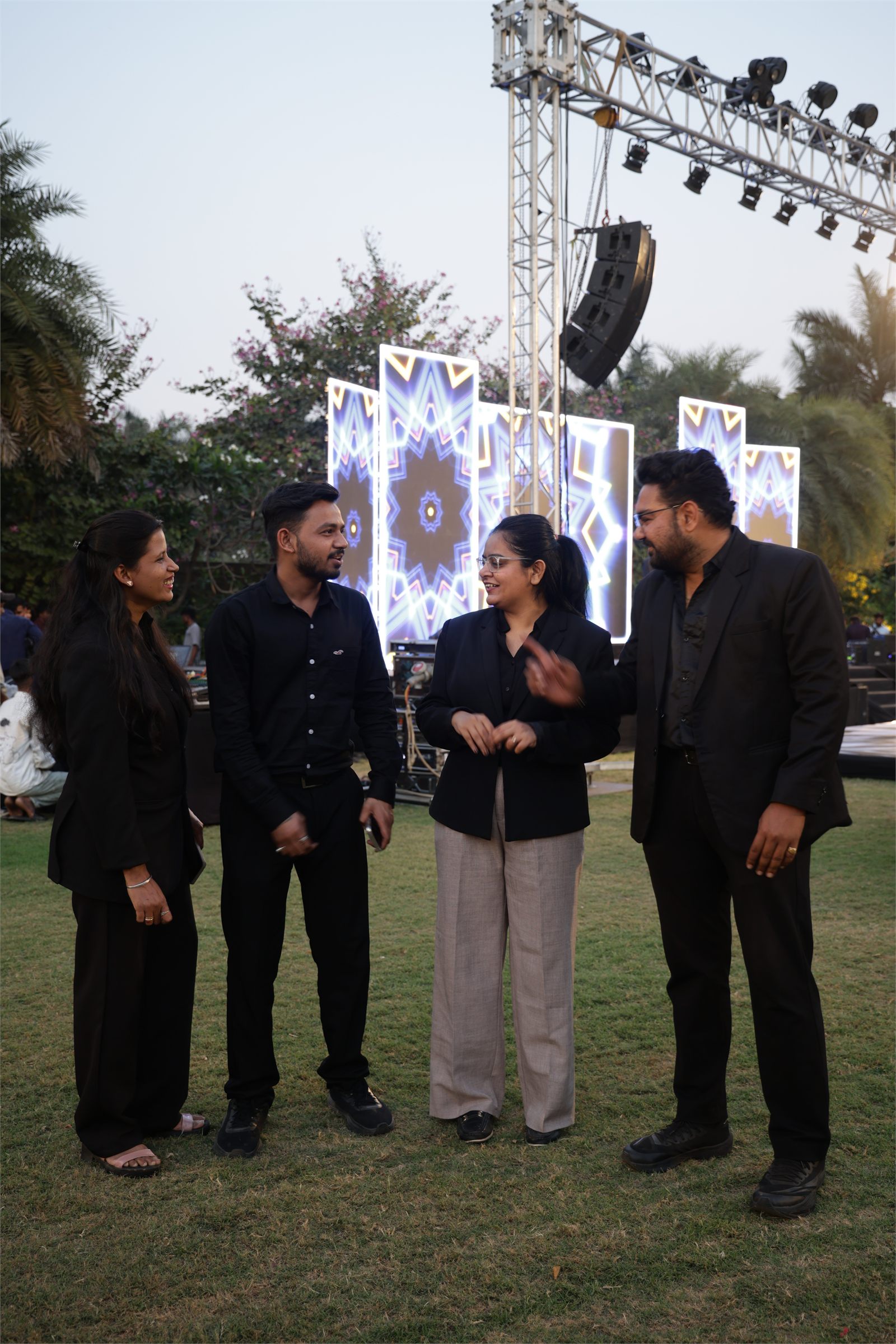 Four Indraa Events crew members talking on a lawn beside a lit LED stage screen at dusk
